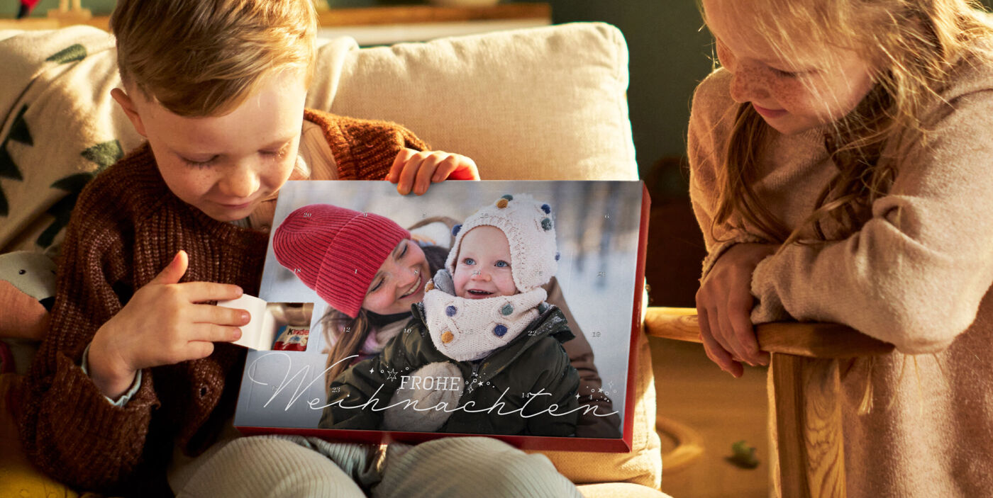 Zwei Kinder öffnen ein Türchen eines CEWE Adventskalenders mit Winterfoto und der Aufschrift „Frohe Weihnachten“ im warmen Licht