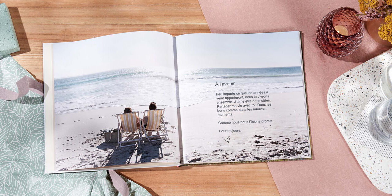Un LIVRE PHOTO CEWE ouvert est posé sur une table. La photo d’un couple au bord de la mer s’étend sur les deux pages. Le titre est « A l’avenir ».