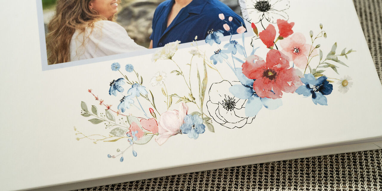 On y voit un gros plan de la couverture du livre d’or. Elle montre une photo du couple de mariés. En dessous, se trouve une décoration florale de mariage sur fond blanc, avec une finition à effet verni.