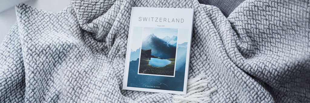 Un LIVRE PHOTO CEWE sur un voyage en Suisse est posé sur une couverture et un coussin à côté d'une plante d'intérieur.