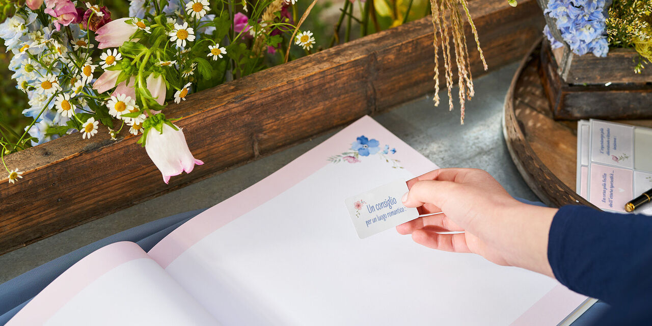 Una mano attacca un foto adesivo con un messaggio di congratulazioni nel libro degli ospiti aperto, che si trova su un tavolo. Accanto al libro degli ospiti ci sono altri foto adesivi. Ci sono anche decorazioni floreali.
