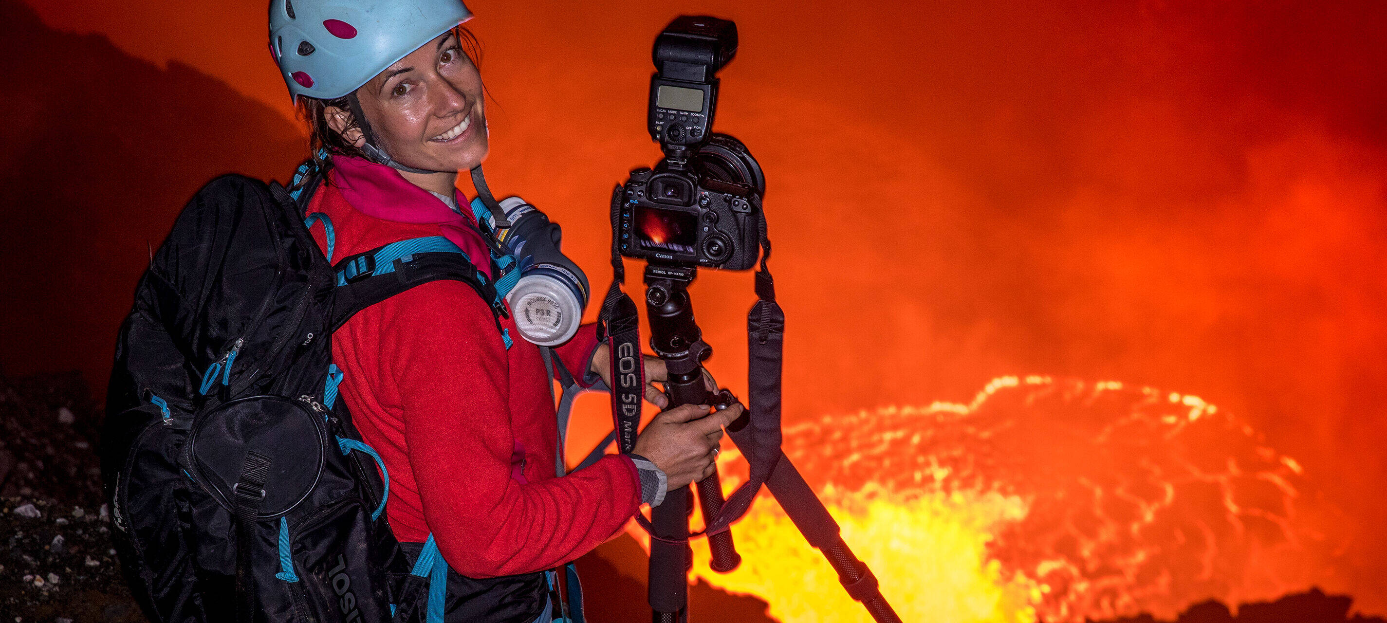 La fotografa di esplorazione Ulla Lohmann è in piedi con casco e zaino sul bordo di un vulcano e sorride alla fotocamera. Accanto a lei c'è una macchina fotografica con treppiede.