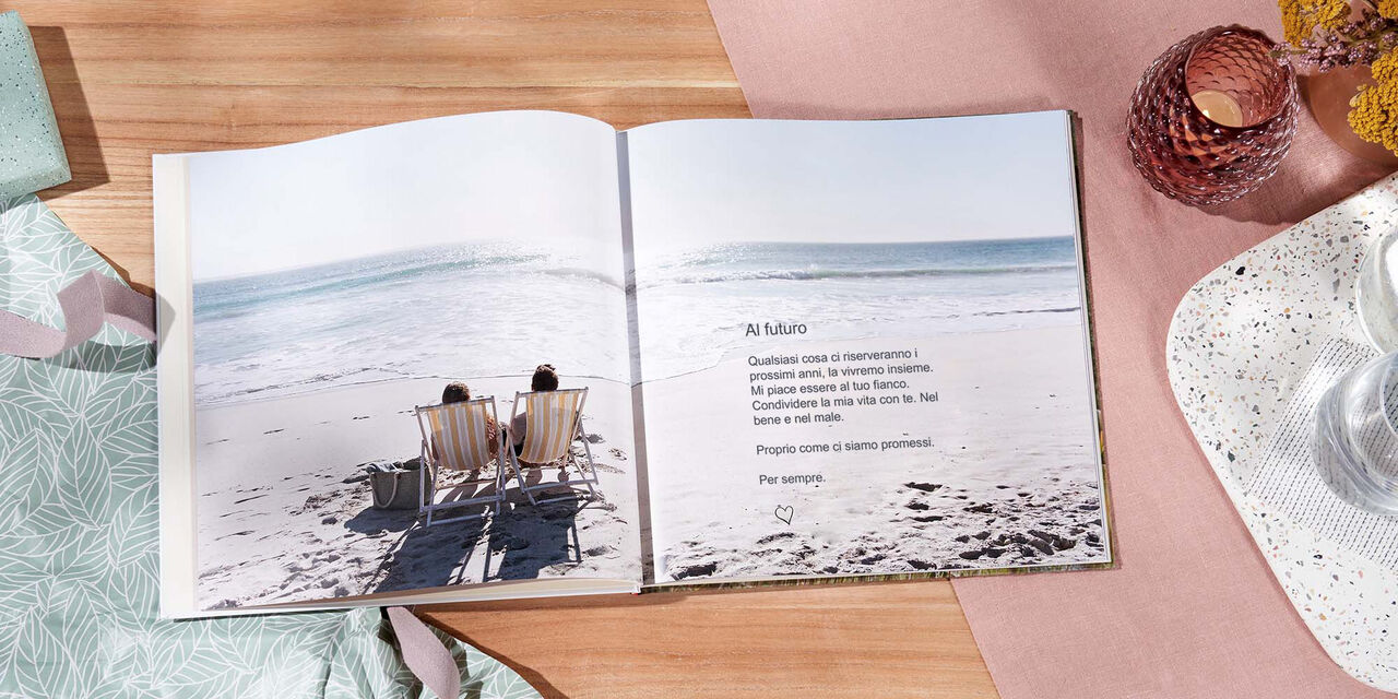 Un FOTOLIBRO CEWE aperto giace su un tavolo. La foto di una coppia in riva al mare si estende su entrambi i lati. La didascalia recita «Al futuro».
